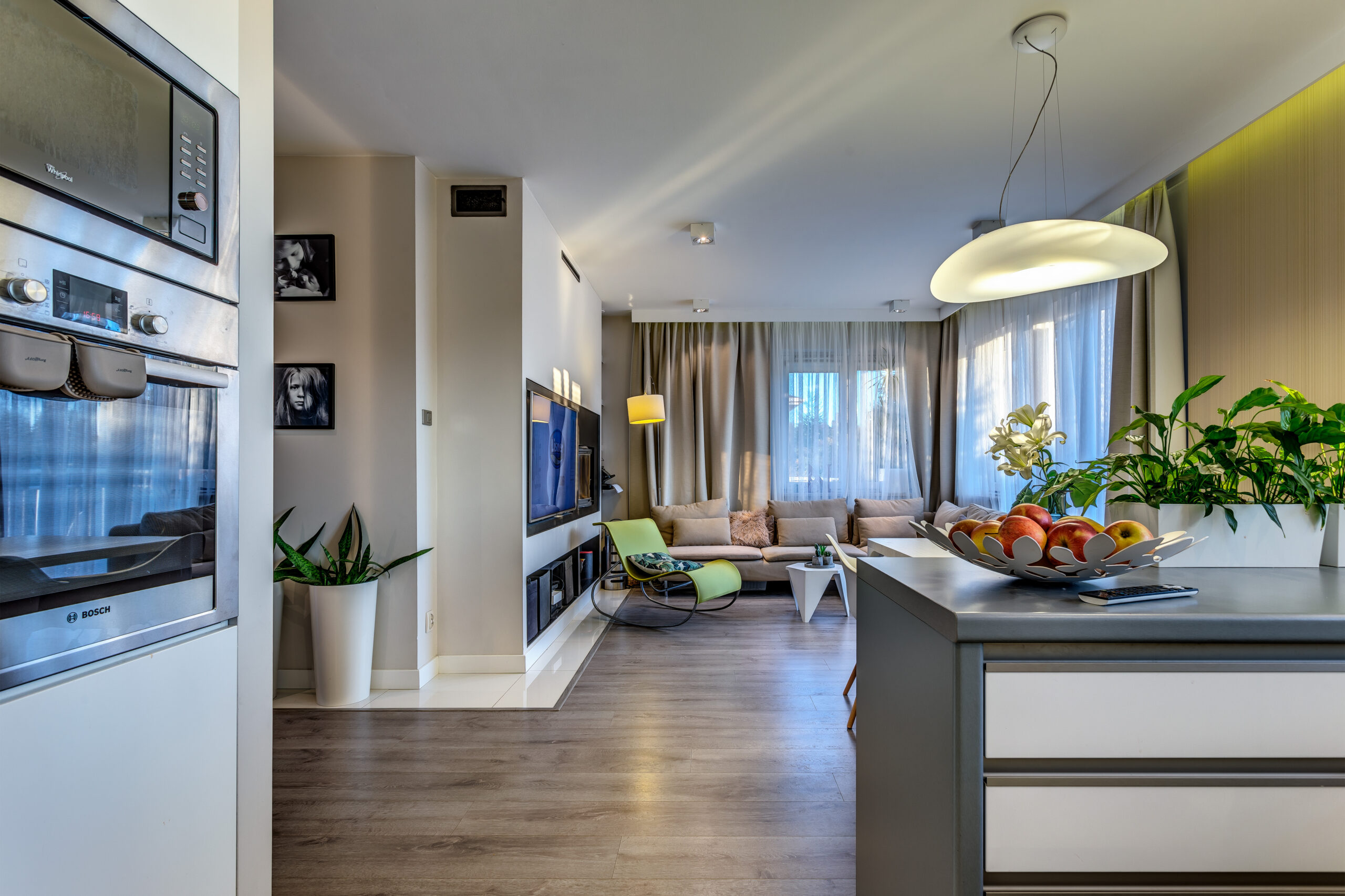 "Przestronne mieszkanie z otwartą kuchnią i salonem. Kuchnia w kształcie litery L, z jasnymi frontami, szarym blatem. Na zdjęciu widoczny jest także przestronny salon, wyposażony w wygodną sofę i fotel. W centralnej części salonu znajduje się elegancki kominek, który dodaje uroku i ciepła temu wnętrzu. Z aneksu kuchennego rozciąga się piękny widok na salon oraz część wypoczynkową. Całość tworzy przytulną przestrzeń, w której można spędzać czas z rodziną i przyjaciółmi."