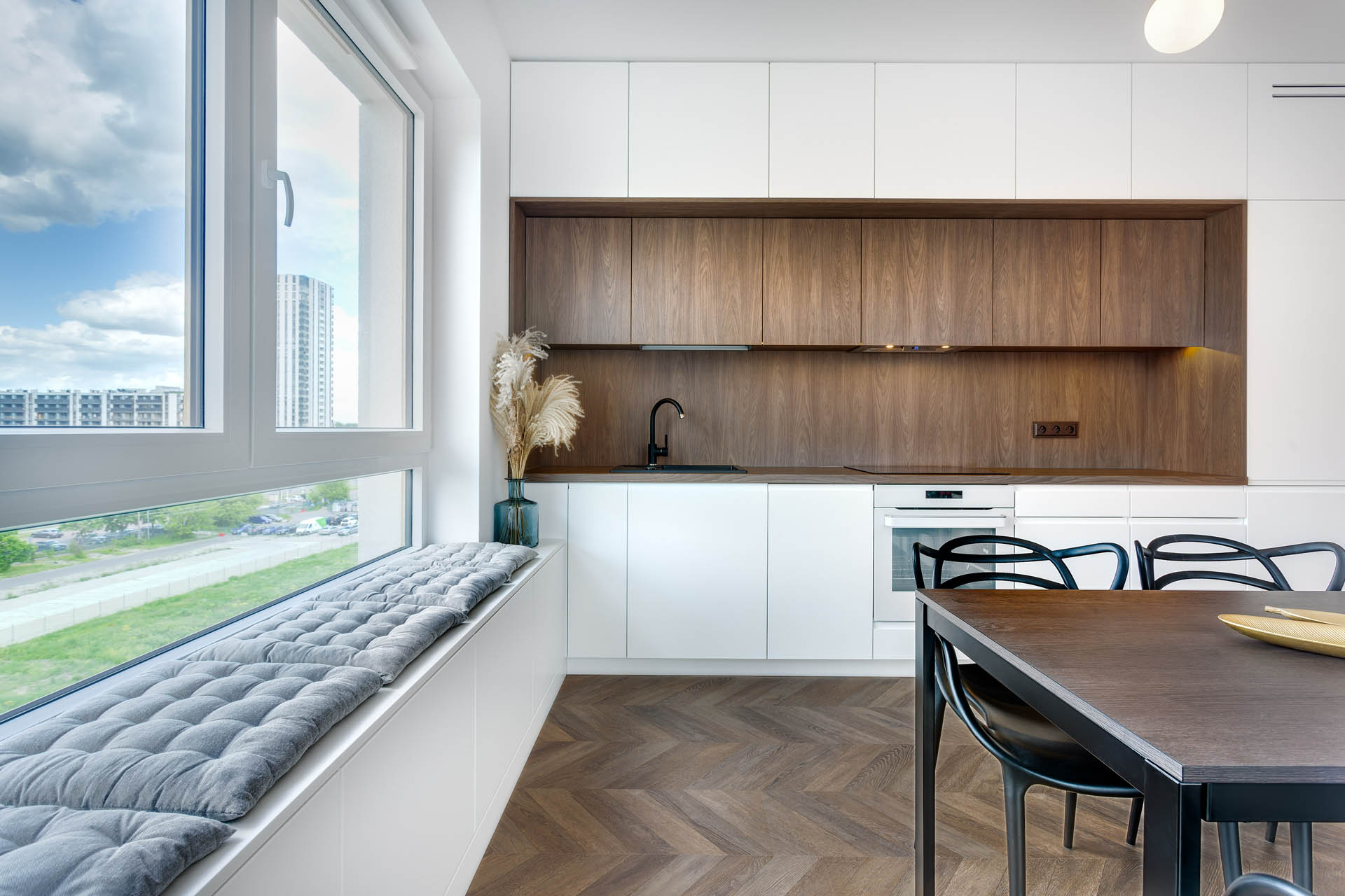 "Przestronne mieszkanie z otwartą kuchnią, salonem oraz jadalnią. Na zdjęciu widoczny jest elegancki, drewniany stół jadalny z krzesłami w nowoczesnym stylu. W kuchni, połączonej z pokojem, znajdują się nowoczesne szafki z frontami w kolorze białym, które nadają pomieszczeniu lekkości i przestronności. Na parapecie okna zaprojektowano wygodne siedzisko, aby odpocząć i podziwiać widok miasta"